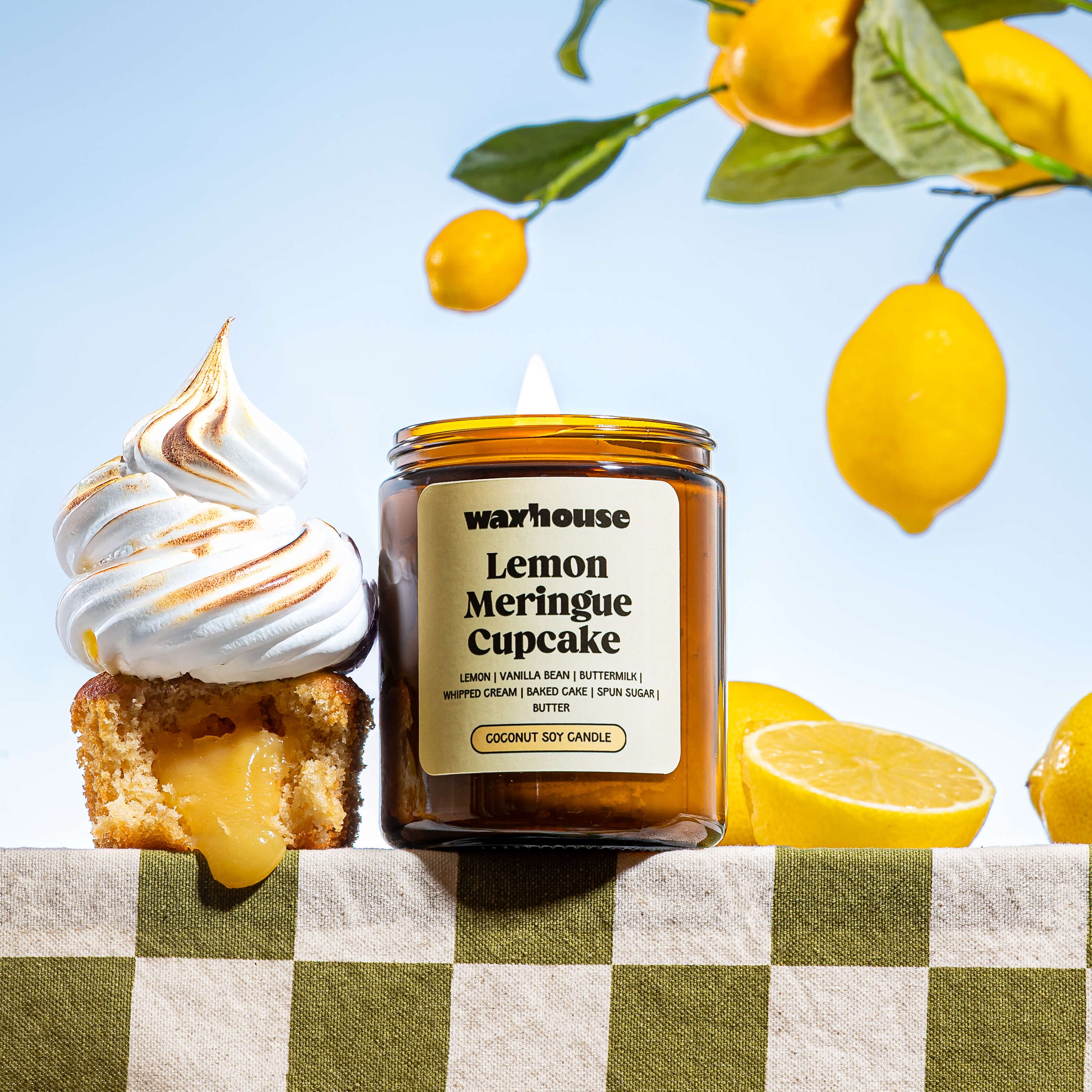 Warhouse Lemon Meringue Cupcake candle with a lemon meringue cupcake and lemons on a checkered tablecloth.